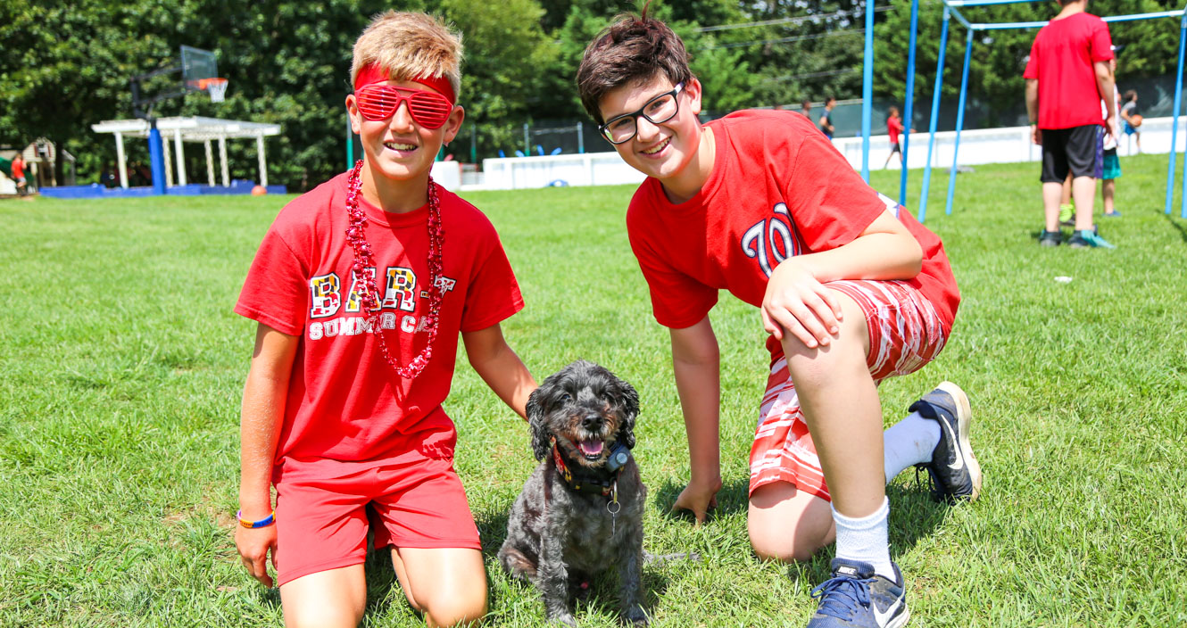 Two campers kneeling down next to a small black dog on the grass