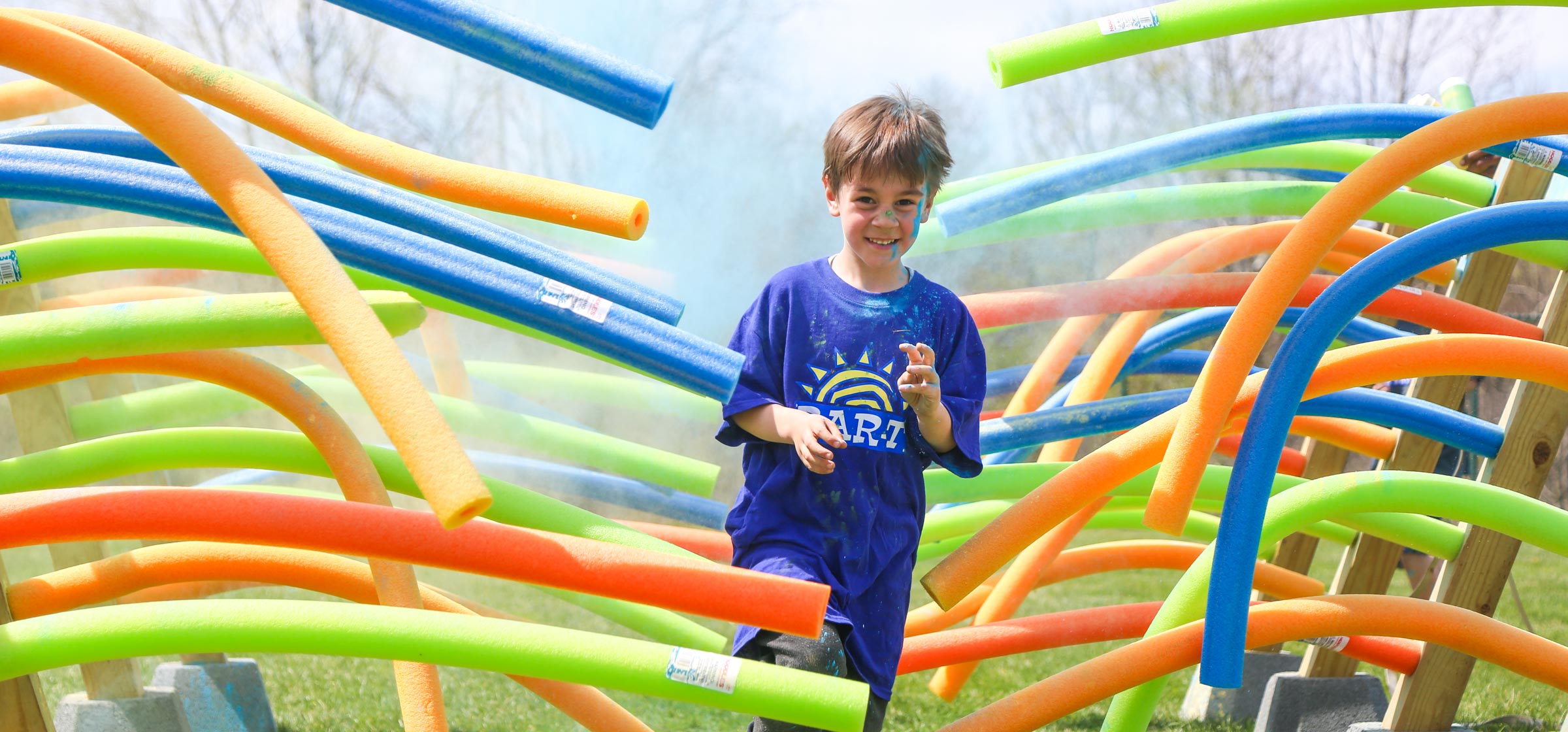 Young boy camper running through the color run course through a bunch of pool noodles