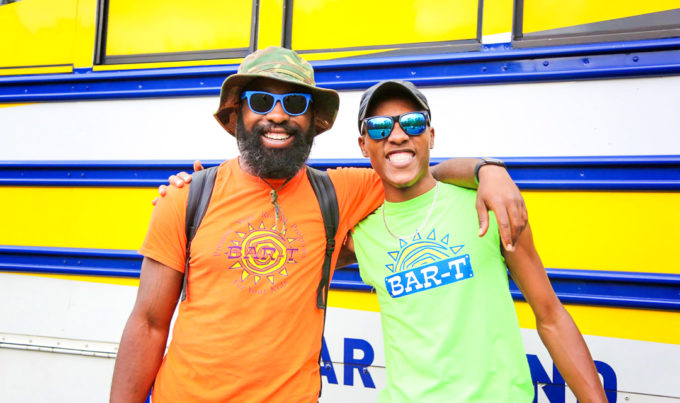 Two counselors with arms around each other wearing sunglasses smiling standing in front of a Bar-T bus