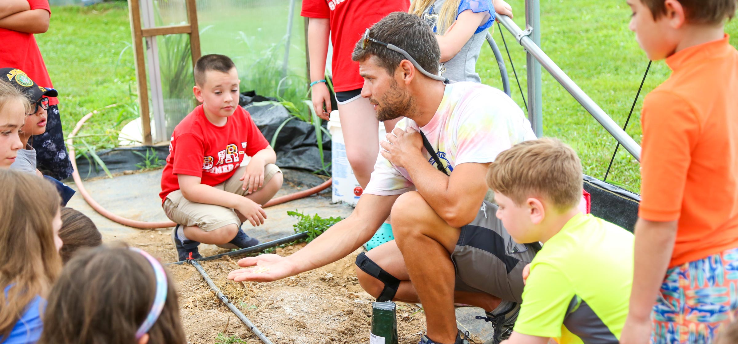 Teacher kneeling with seeds in his palm showing students gathered around in a greenhouse