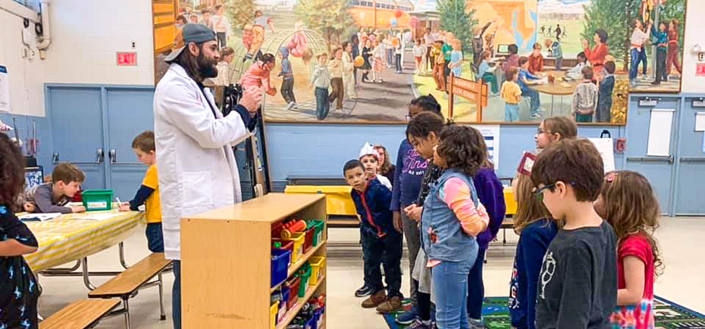 Professor Ross demonstrating something to a group of kids lined up in front of him in a classroom